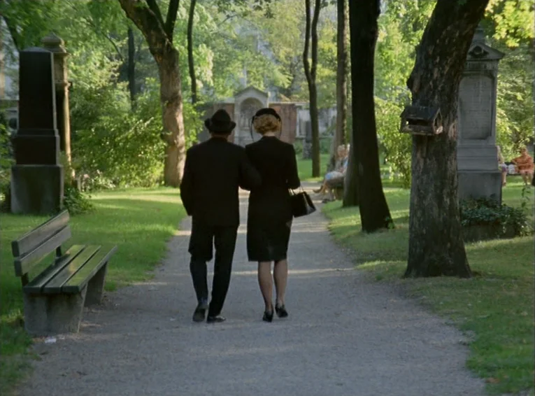 The Merchant Of The Four Seasons (1971) movie still: fullbody — A gravel path runs between rows of headstones under a canopy of mature trees. A man…