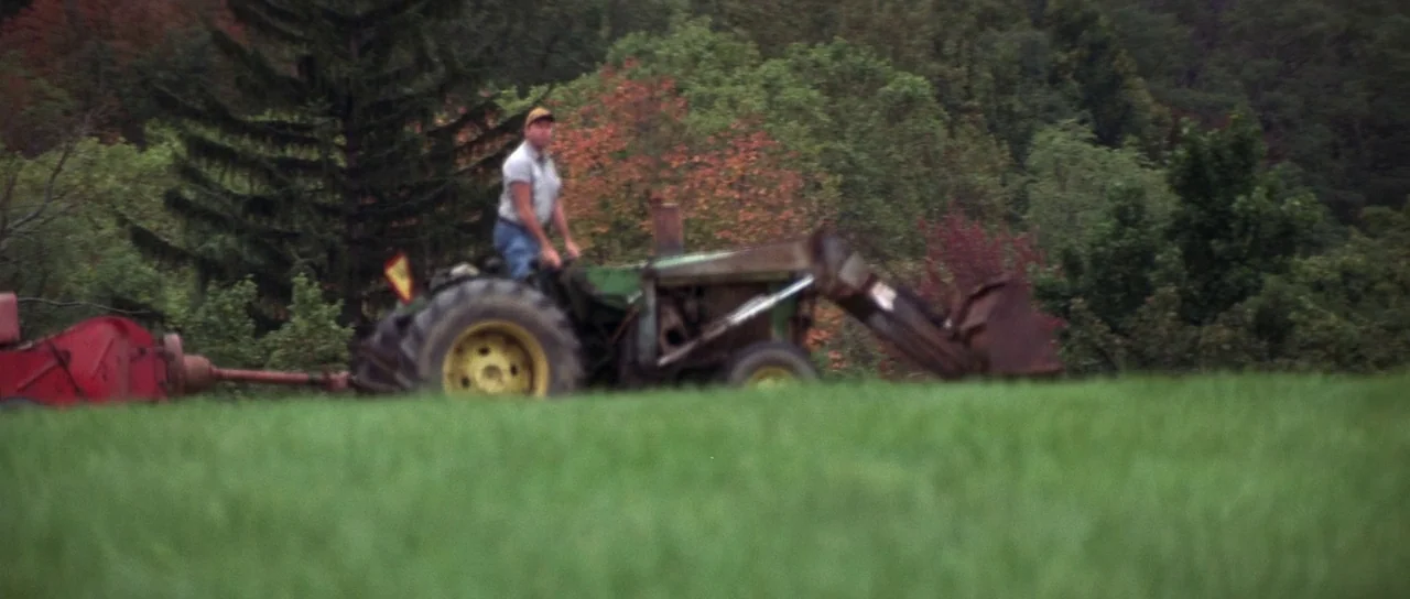 The Thomas Crown Affair (1999) movie still: wide — A man drives a vintage green tractor across a large, lush green field. Dense woodland with…