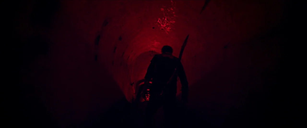 Mandy (2018) movie still: medium — A dark, curved stone tunnel is lit by a harsh red light source. A lone figure walks away from the camera through…