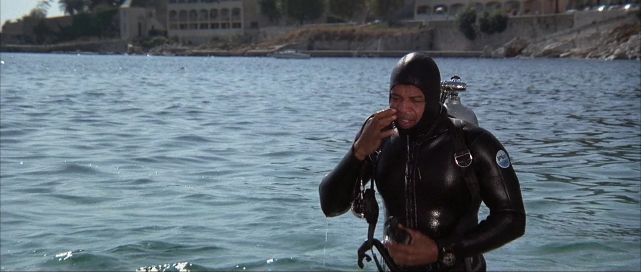 Never Say Never Again (1983) movie still: medium — A man stands in calm shallow water near a rocky coastline and low buildings. The diver holds his…