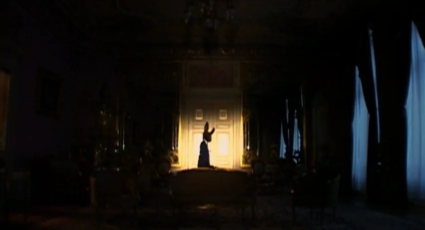 Inland Empire (2006) movie still: wide — A dark, ornate room with antique furniture is lit by light from double doors. A figure wearing a rabbit…