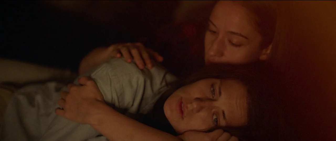 Nightsiren (2022) movie still: closeup — A dimly lit room at night features two people lying close together in shadow. One woman rests her head on…