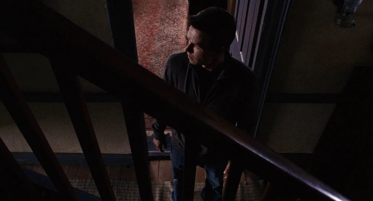 The Happening (2008) movie still: medium — A dark, dimly lit staircase landing features a red patterned rug and wood railings. A man stands on the…