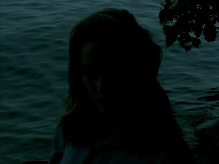 Helas Pour Moi (1993) movie still: closeup — A woman stands near dark, rippling water under low light conditions at night. Her face is obscured by…