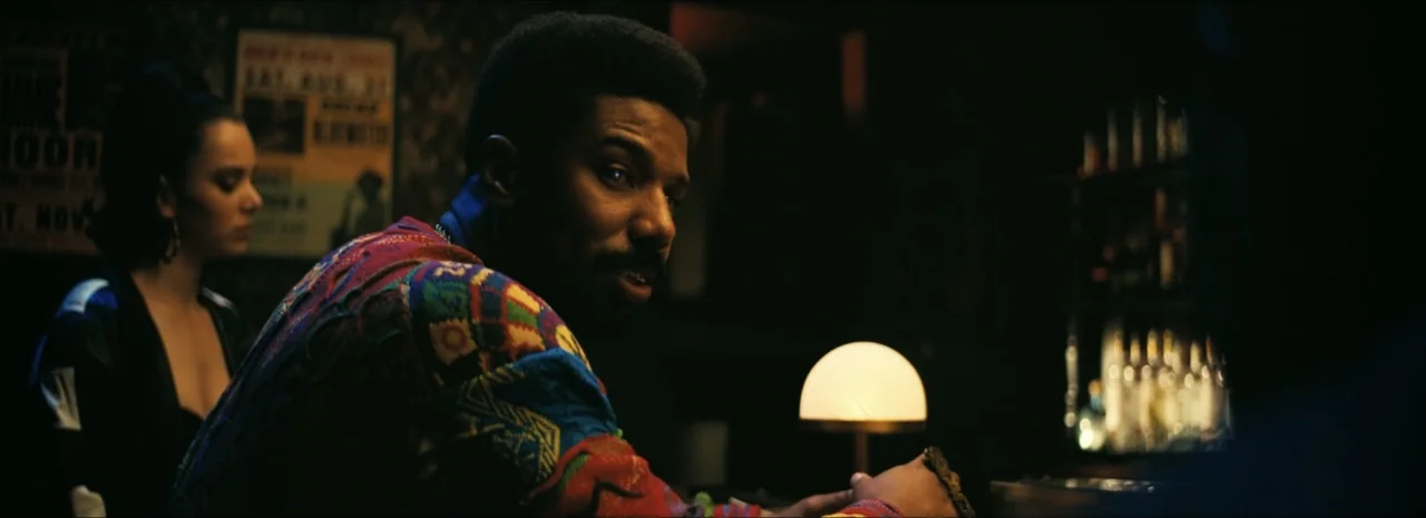 Sinners (2025) movie still: medium — A dim bar interior features a man in a patterned sweater sitting at the counter. Beside him, a woman sits in…
