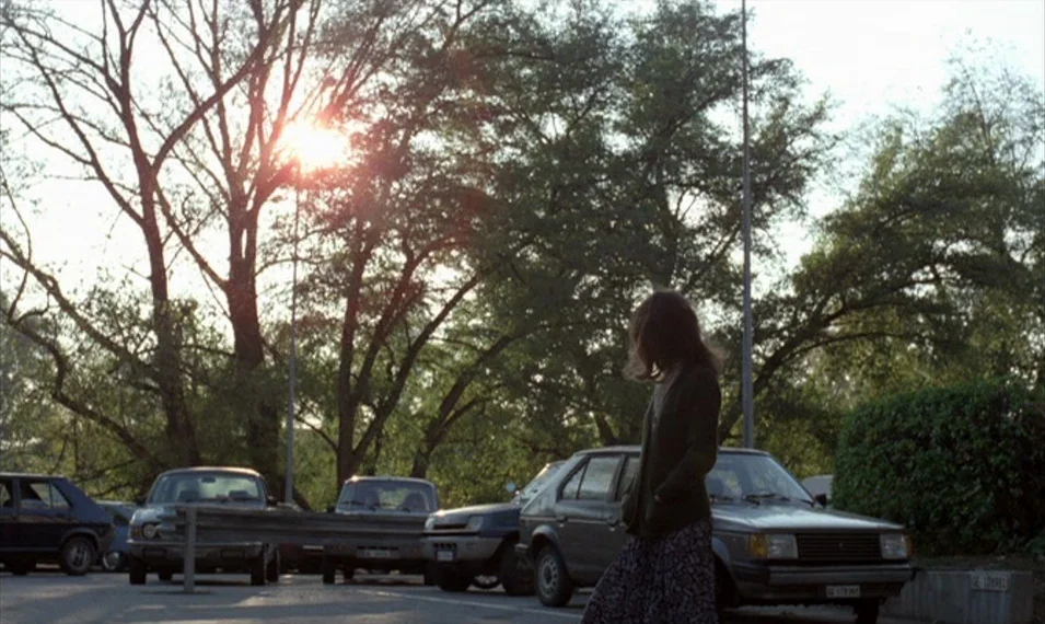 Every Man for Himself (1980) movie still: medium — An asphalt parking lot features several 1980s-era cars parked in front of mature trees. A woman…