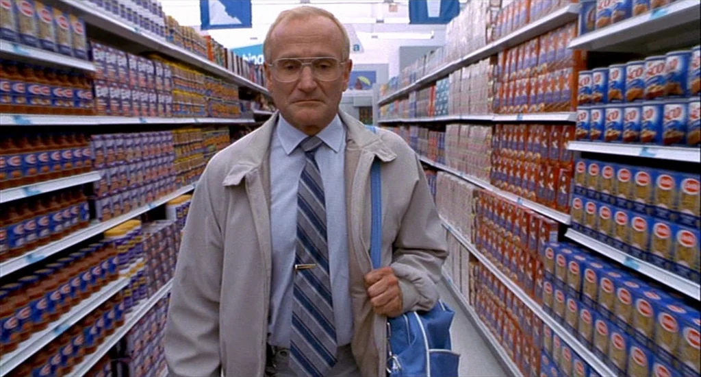 One Hour Photo (2002) movie still: medium — An aisle in a retail store is filled with rows of stacked food products. An elderly man walks toward the…