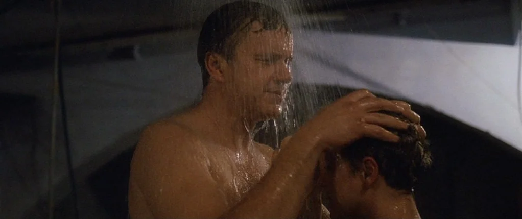 Code 46 movie still: medium — A dark tiled shower stall is illuminated by streams of water. A shirtless man stands close to another person, placing…