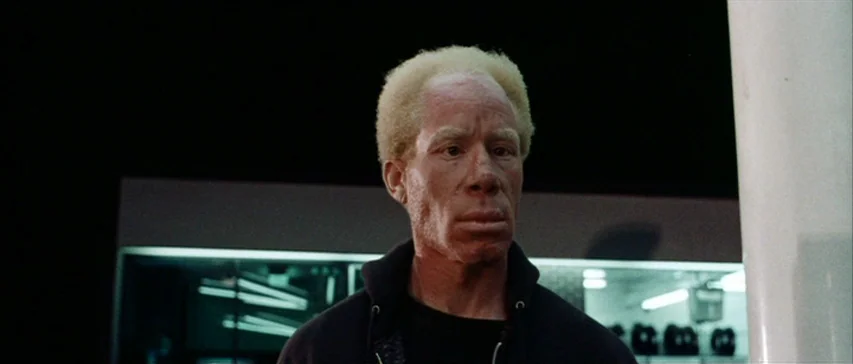 Messiah of Evil (1973) movie still: medium — A man stands outside a gas station at night under bright artificial light. He looks away from the…