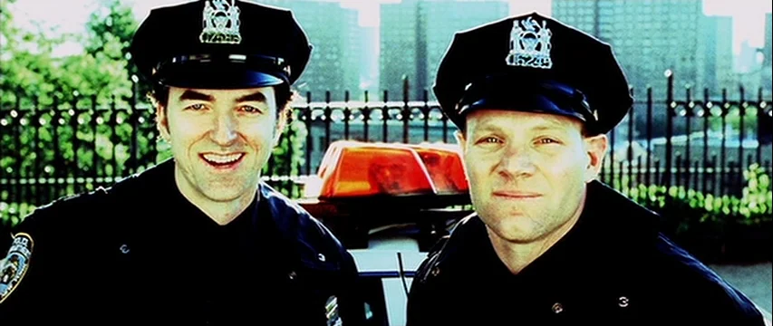 25th Hour (2002) movie still: medium — Two police officers stand in front of a patrol car near a metal fence. The officers face the camera while…