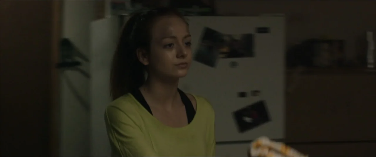 Captive (2020) movie still: medium — A dark, dimly lit kitchen interior features a white refrigerator adorned with several magnets. A young woman…
