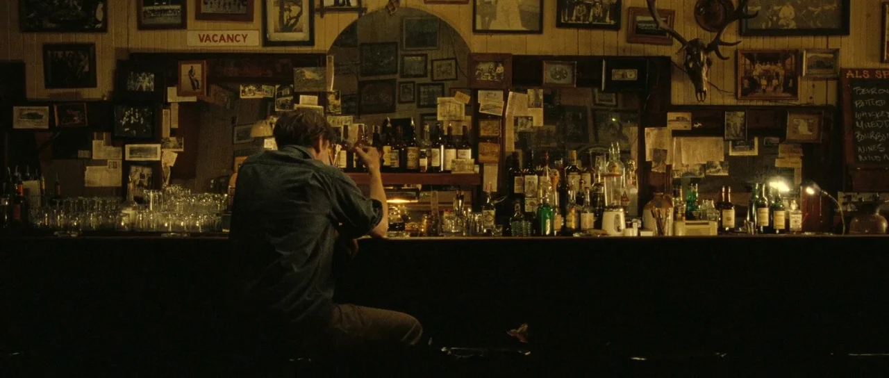 On The Road (2012) movie still: medium — A dimly lit bar wall is covered in numerous framed photos and a skull mount. A man in a blue shirt sits…