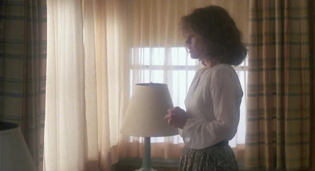 Coming Home (1978) movie still: medium — A woman stands in front of a window covered by layered cream curtains. She keeps her hands clasped together…