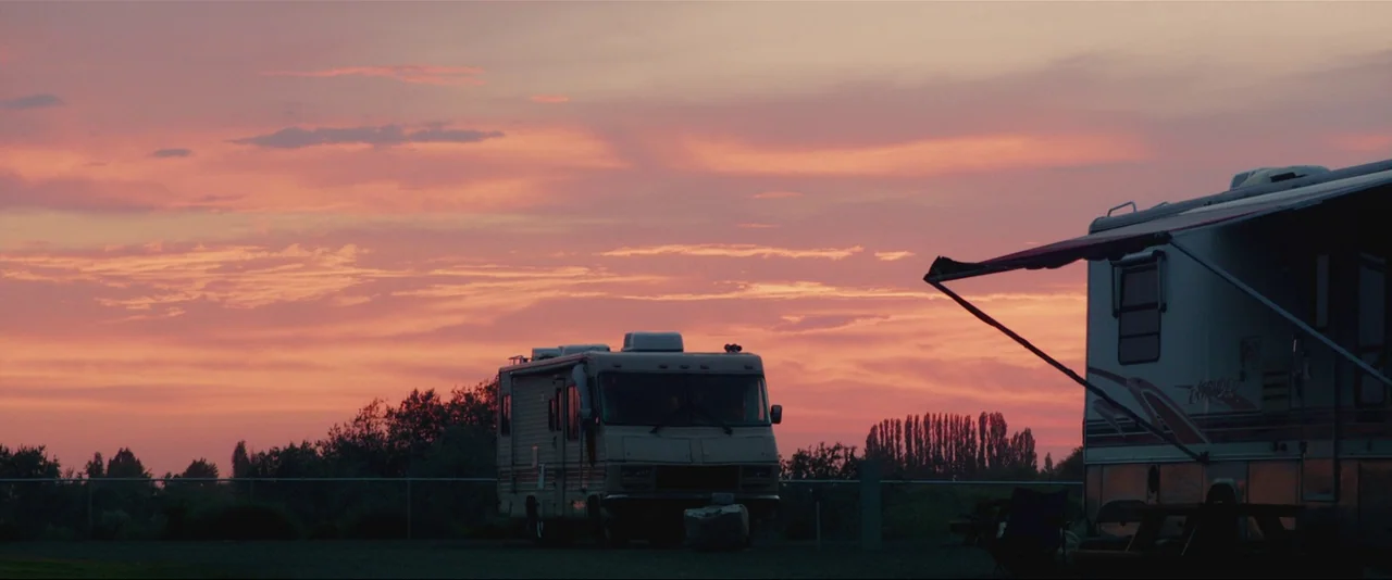 Captain Fantastic (2016) movie still: establishing — A quiet RV park at sunset features two parked motorhomes against a pink horizon. The sky is…