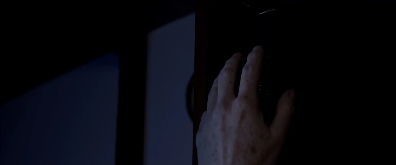 Mermaid Down (2019) movie still: closeup — A dimly lit interior space features a dark wall area and an obscured background. An aged hand extends…