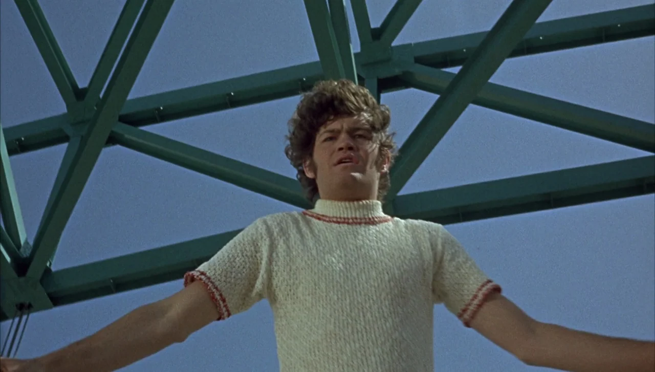 Head (1968) movie still: medium — A man stands in front of a teal painted steel truss structure outside. The subject is positioned centrally in the…