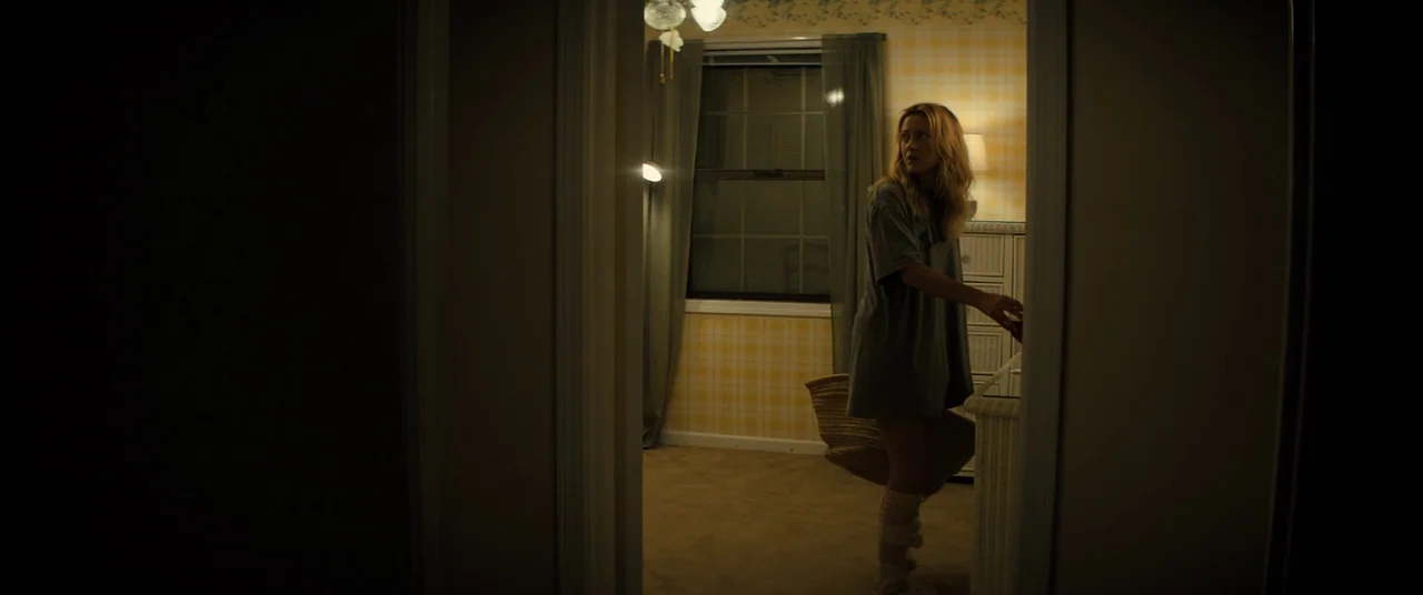 Brightburn (2019) movie still: medium — A dimly lit room with plaid wallpaper and a wooden baby crib. A young woman stands inside the room reaching…