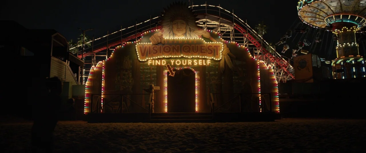 Us (2019) movie still: wide — A dark, sandy amusement park at night features a brightly lit carnival funhouse facade. The building is decorated with…