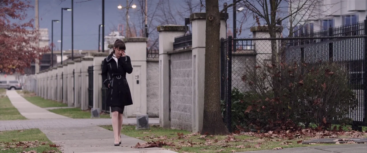 American Mary (2012) movie still: fullbody — A woman walks down a sidewalk lined by a concrete wall and metal fencing. The environment is a grey…