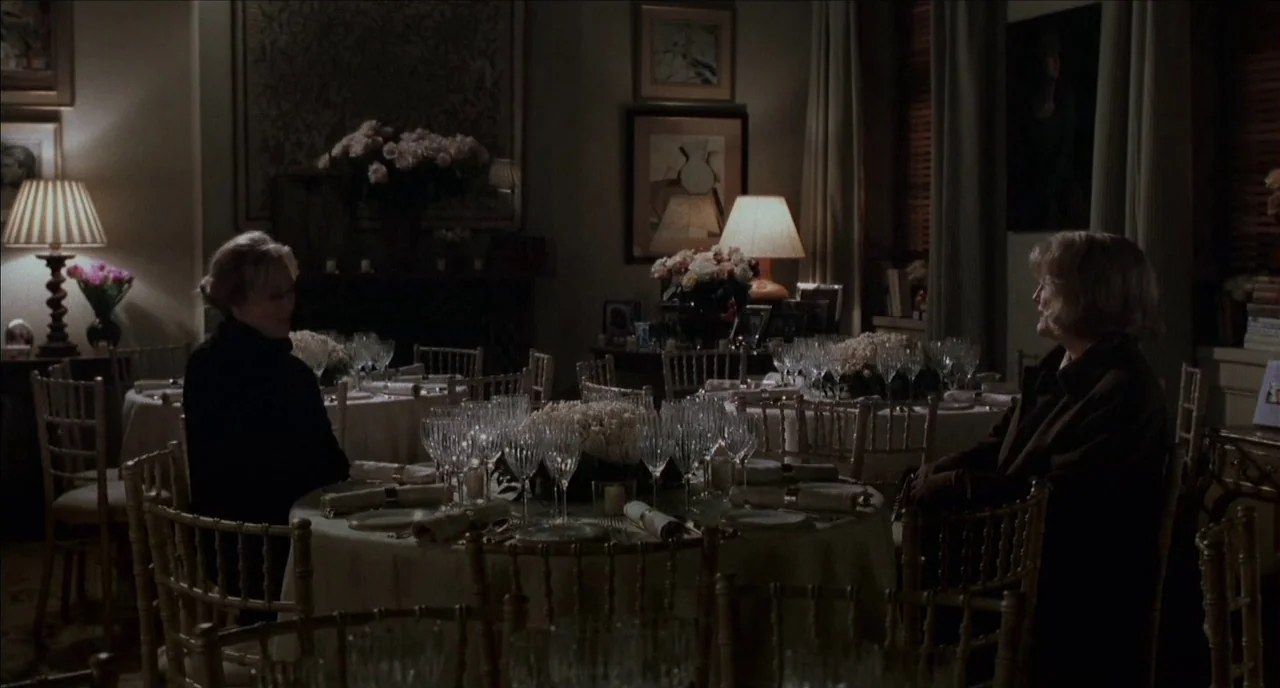 The Hours (2002) movie still: wide — A dimly lit room contains multiple round tables draped in pale white tablecloths. Two women sit across from one…