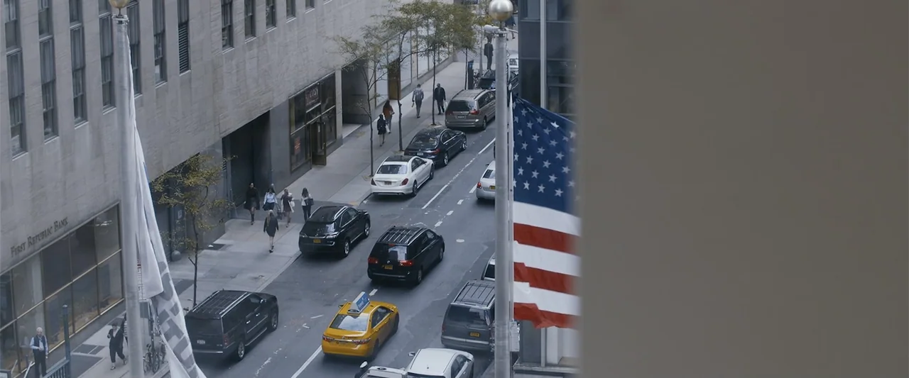 The Kleptocrats (2018) movie still: wide — A dense urban city street is viewed from an elevated vantage point above. Several cars and a yellow taxi…