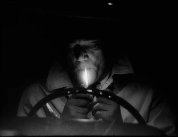 Alphaville (1965) movie still: medium — A dark car interior at night with a steering wheel in the foreground. A man holds a lit cigarette lighter to…