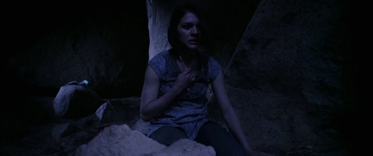 Here Comes The Devil (2012) movie still: medium — A dark, cramped space surrounded by large jagged rock formations and sandy ground. A young woman…