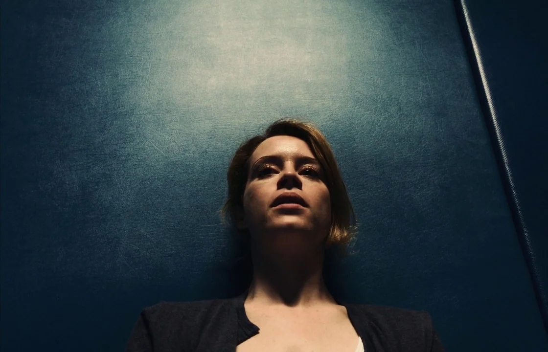 Unsane (2018) movie still: closeup — A dimly lit, narrow metal wall interior with a deep blue textured surface. A woman stands against the wall…