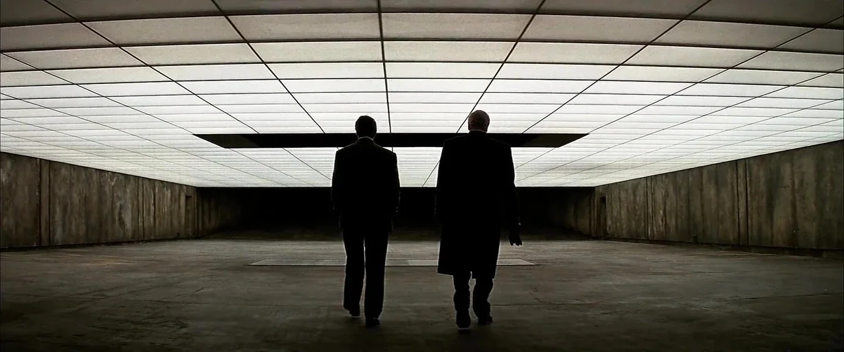The Dark Knight (2008) movie still: wide — A vast, minimalist concrete room features a ceiling entirely composed of glowing square light panels. Two…