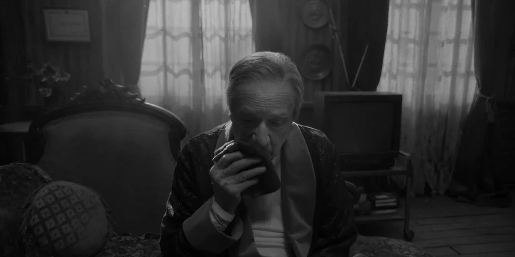 El Conde (2023) movie still: medium — A dimly lit room contains an ornate sofa and a television set on a cart. An elderly man in a robe sits on the…