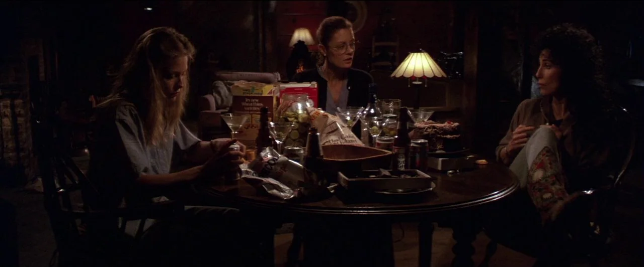 The Witches of Eastwick (1987) movie still: medium — A dimly lit room features a round wooden table crowded with snacks and drinks. Three women sit…
