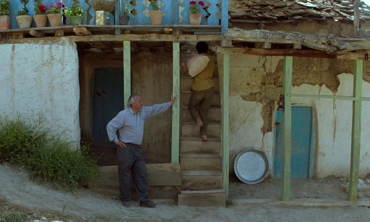 Through The Olive Trees (1994) movie still: fullbody — A rural dwelling made of mud bricks has a blue balcony and outdoor stairs. One man stands by…
