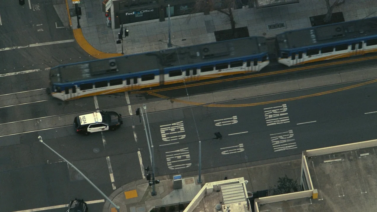 One Battle After Another (2025) movie still: establishing — An aerial view captures an urban intersection featuring a light rail train and vehicle.…