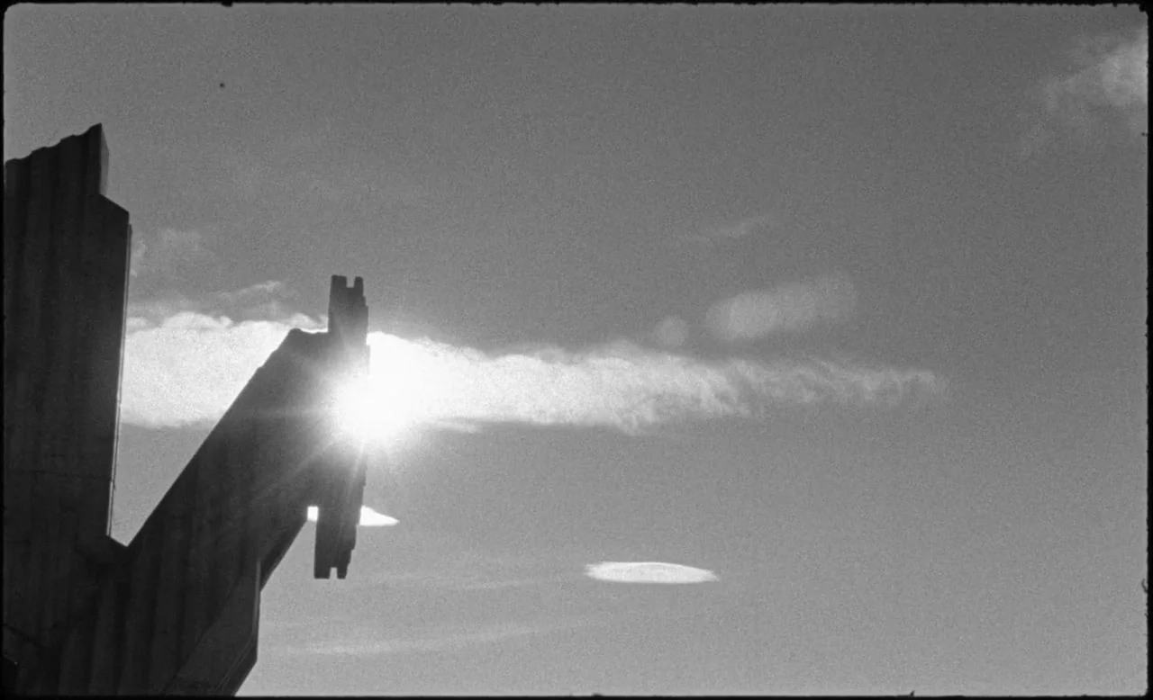 Last and First Men (2020) movie still: establishing — A dark concrete monument rises into a bright, slightly clouded sky at daytime. The sun is…