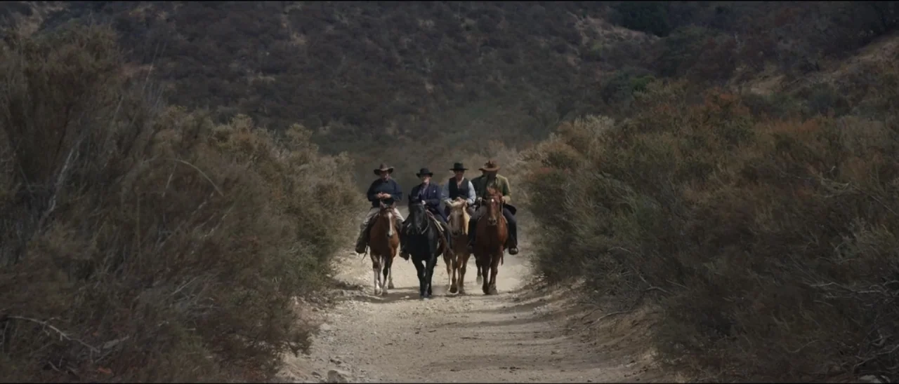 Dead for a Dollar (2022) movie still: wide — A dry, dusty mountain trail is flanked by dense, scrubby desert vegetation. Four men on horseback ride…