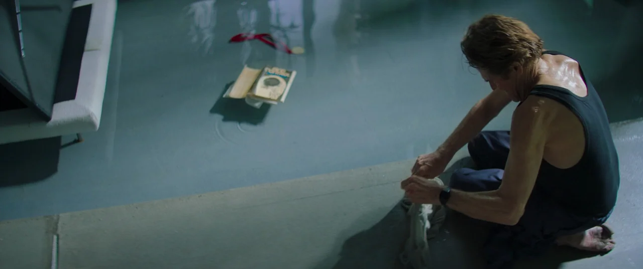 Inside (2023) (2023) movie still: medium — A man kneels on the edge of a tiled basin filled with water. A book and a small medal float on the water…