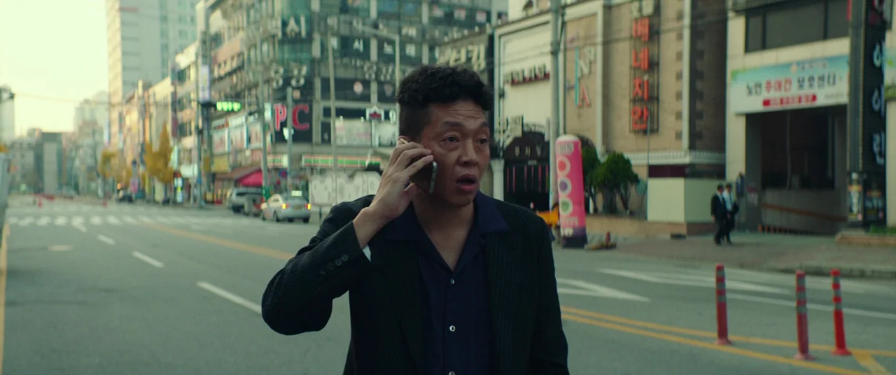Beasts Clawing At Straws (2020) movie still: medium — An empty urban road lined with multi-story buildings featuring prominent neon and printed…