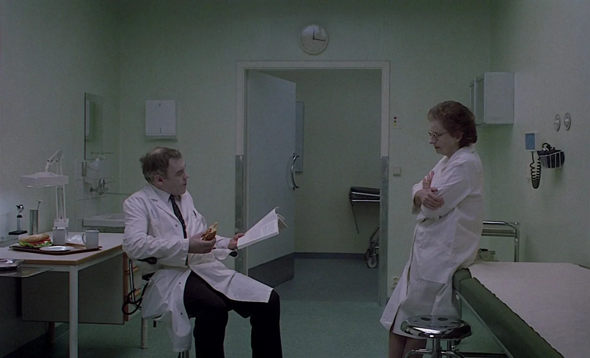 Songs from the Second Floor (2000) movie still: wide — A clinical examination room has pale green walls, medical equipment, and an open doorway. Two…