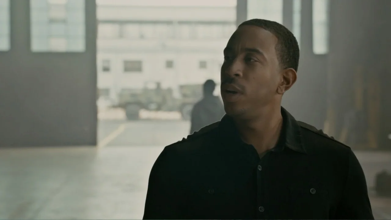 Furious 7 (2015) movie still: medium — A man stands inside a large industrial warehouse with a bright open doorway nearby. He is speaking while…