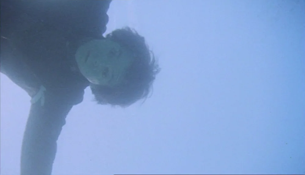 Harold and Maude (1971) movie still: medium — A person floats underwater in a body of blue open water. The surface ripples are visible above the…