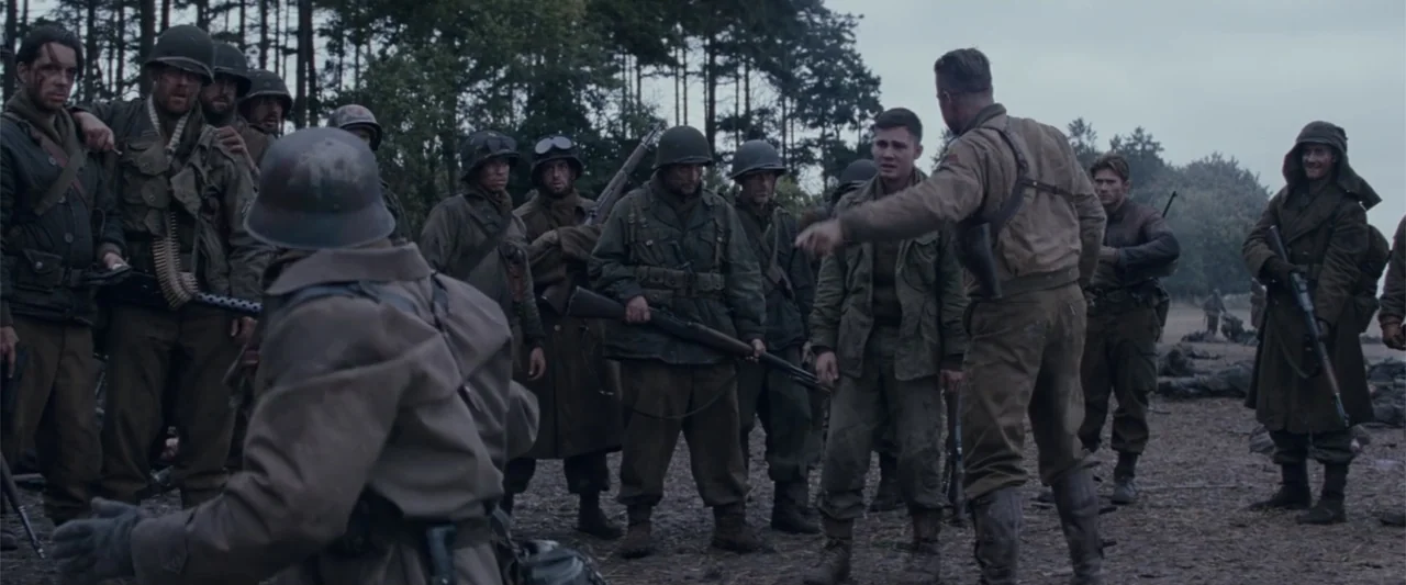 Fury (2014) movie still: wide — A group of soldiers stands in a forest clearing surrounded by tall pine trees. One soldier in a tan field jacket…