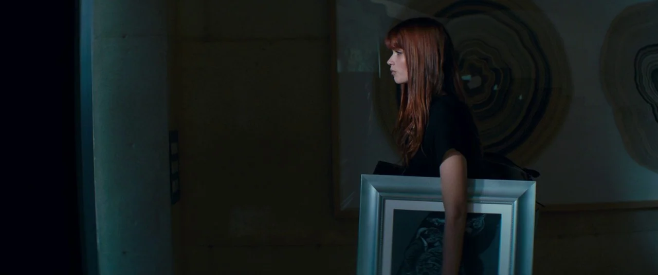 Elizabeth Harvest movie still: medium — A dimly lit indoor space with circular abstract artwork mounted on the walls. A young woman with auburn hair…