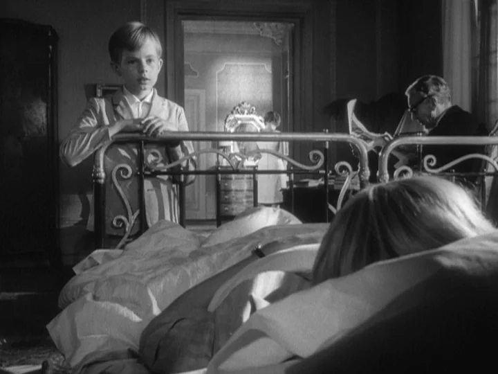The Silence (1963) movie still: medium — A quiet room contains a metal-framed bed where someone is resting under linens. A boy stands at the foot of…