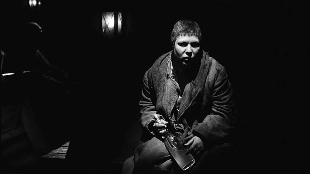 The Role (2013) movie still: medium — A dark, cramped interior space illuminated only by a dim overhead light source. A man in a heavy coat sits…