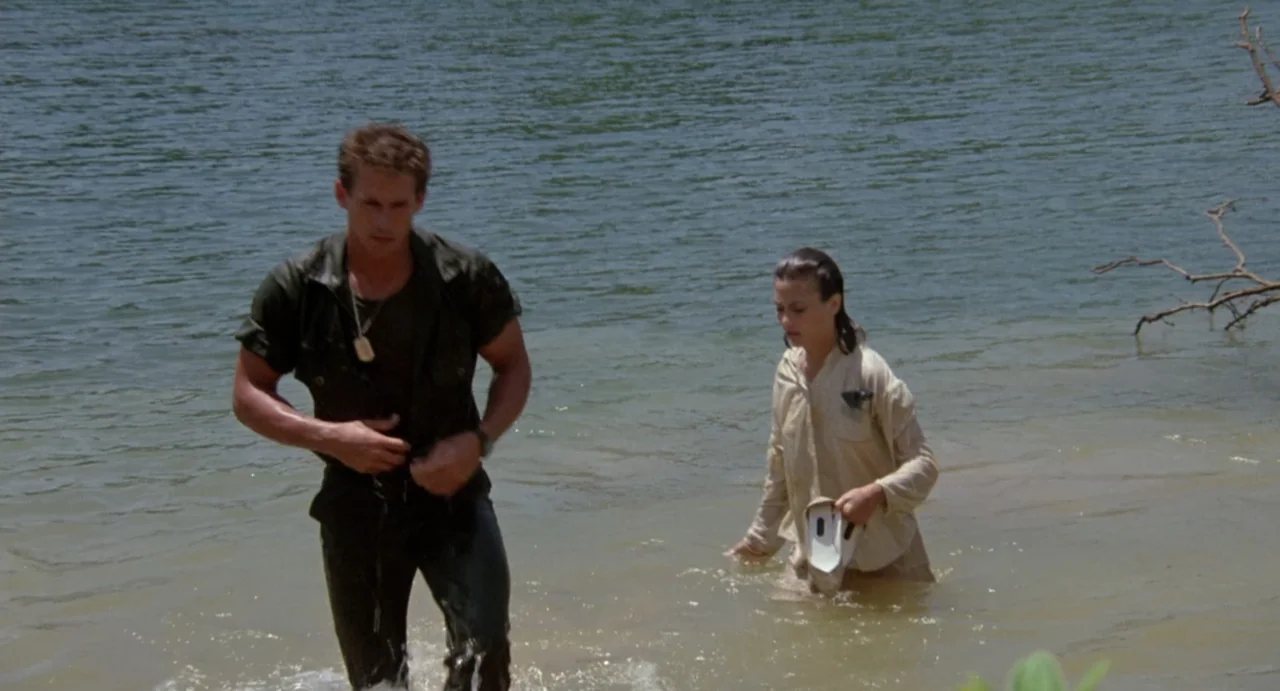 American Ninja (1985) movie still: fullbody — A man and a woman walk through shallow lake water toward the sandy bank. They are wet from the water…