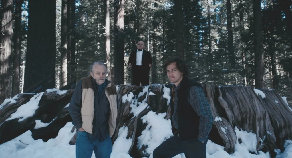 My Son, My Son, What Have Ye Done? (2009) movie still: fullbody — A snowy woodland setting features a massive, weathered tree stump surrounded by…