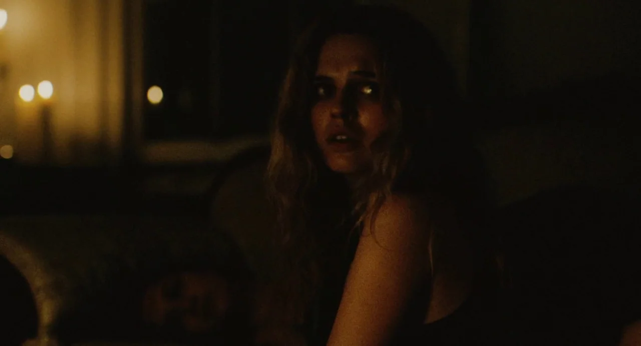 The Giant (2019) movie still: medium — A dark bedroom interior illuminated by soft, warm light from unseen practical lamps. A young woman with long…