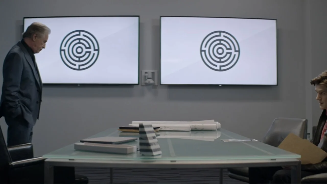 The Collective (2023) movie still: medium — A modern, minimalist meeting room features two large screens displaying matching circular maze logos. An…