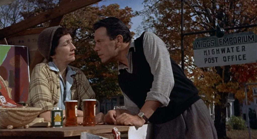 The Trouble With Harry (1955) movie still: medium — An outdoor market stall stands before autumn trees with a wooden sign board nearby. A man in a…