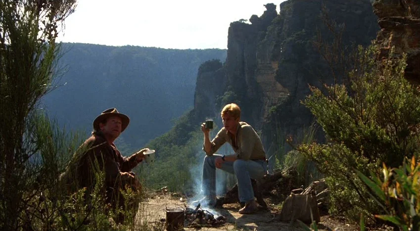 The Coca-Cola Kid (1985) movie still: wide — A rugged cliff edge overlooks a vast valley featuring sheer rock faces and vegetation. Two men sit on…
